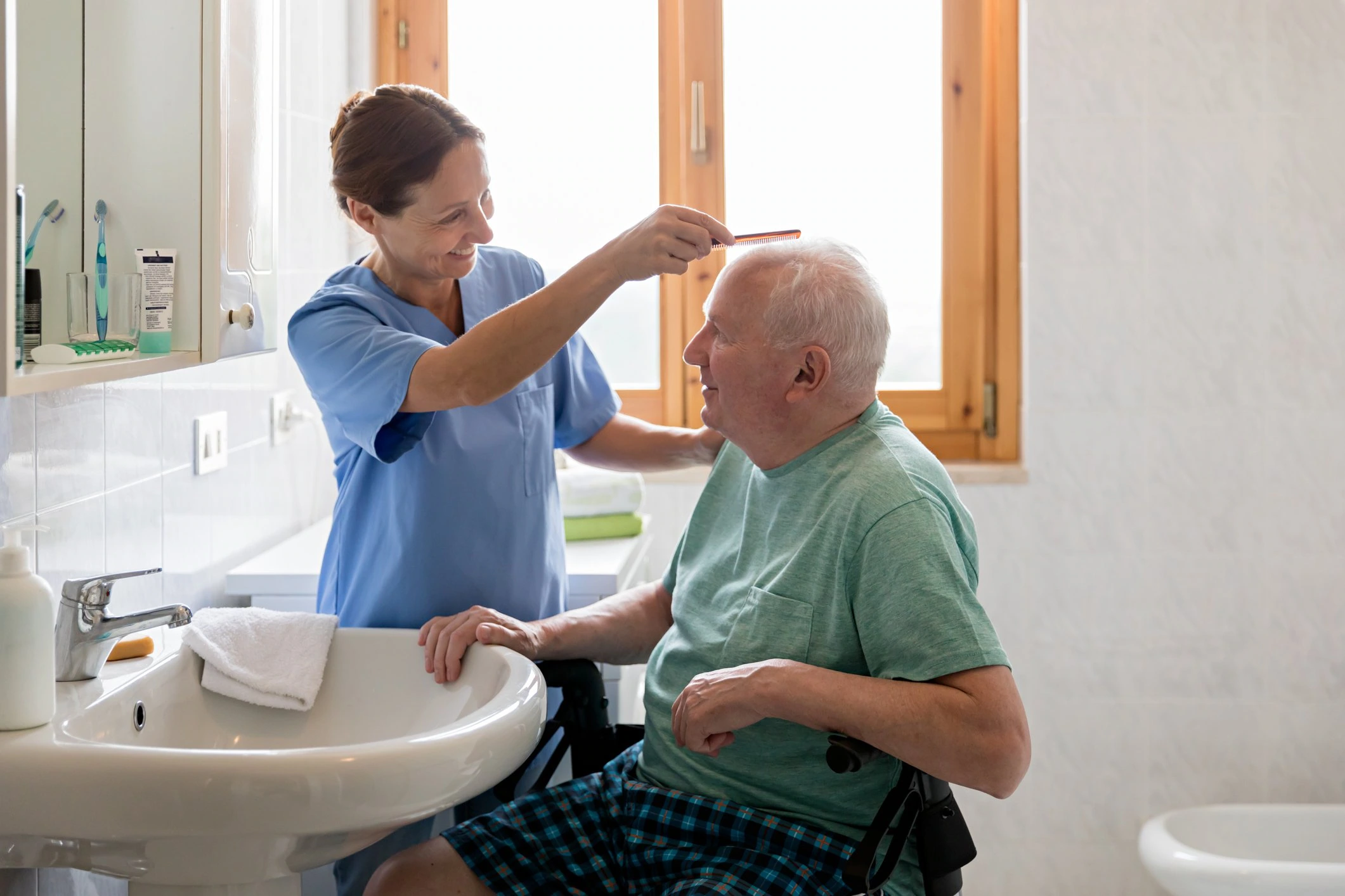 A direct care worker combing an older gentleman's hair in front of a bathroom sink.