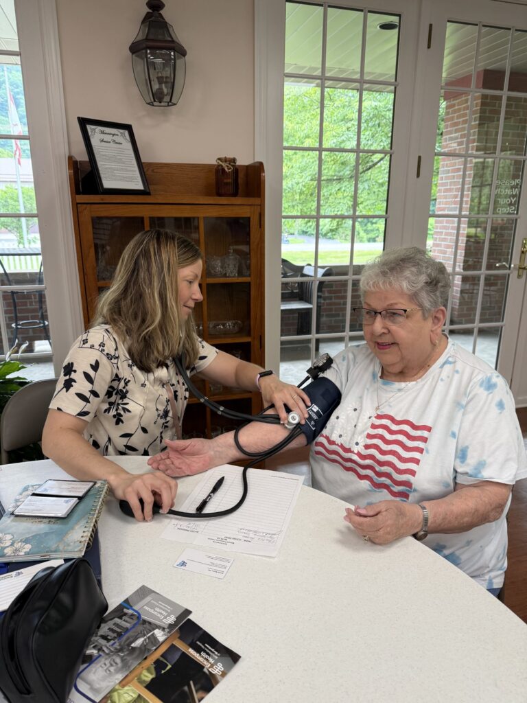 Senior woman wearing a white tee with American flag on front getting her blood pressure taken by a woman in a tan and black blouse with long light brown hair